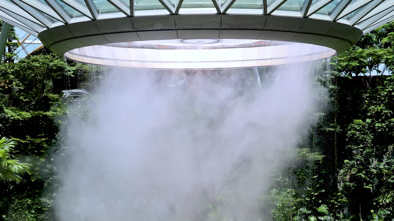 A dramatic indoor waterfall pours from a circular skylight into a verdant, glass-roofed conservatory. Natural daylight illuminates dense tropical foliage as mist fills the air