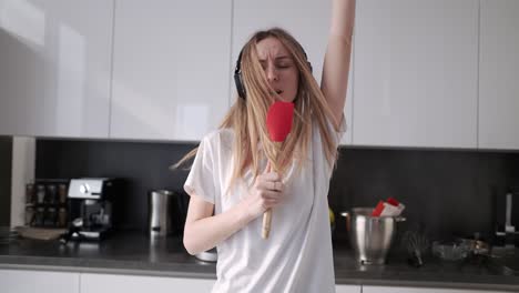 Mujer-Joven-En-Auriculares-Bailando-Y-Fingiendo-Cantar-En-La-Cocina-En-Casa