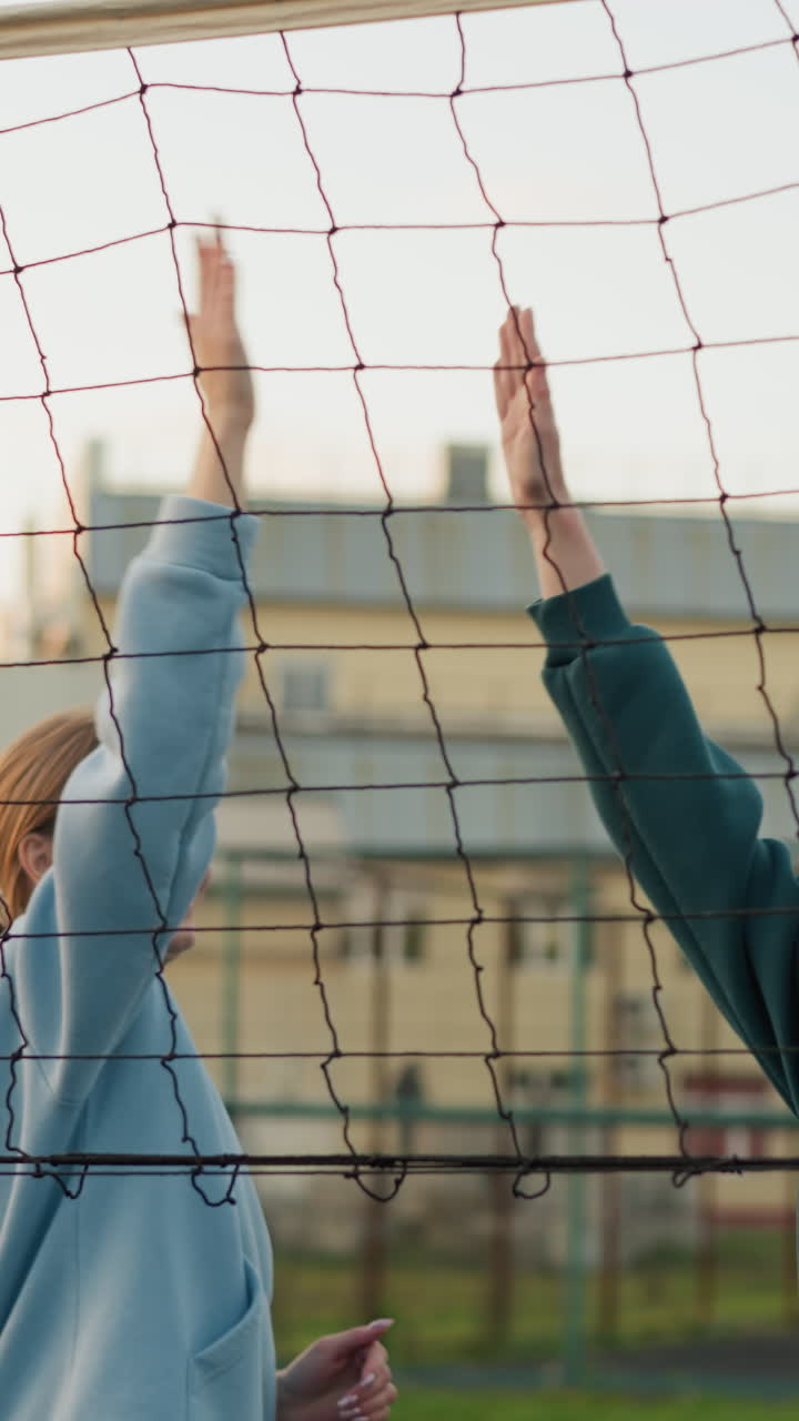 초록색과 파란색 후디를 입은 팀 동료들이 함께 점프하고, 배구를 다시 네트로 보내며, 손을 잡고 웃으며 승리를 축하하며, 배경에는 건물들이 있습니다.