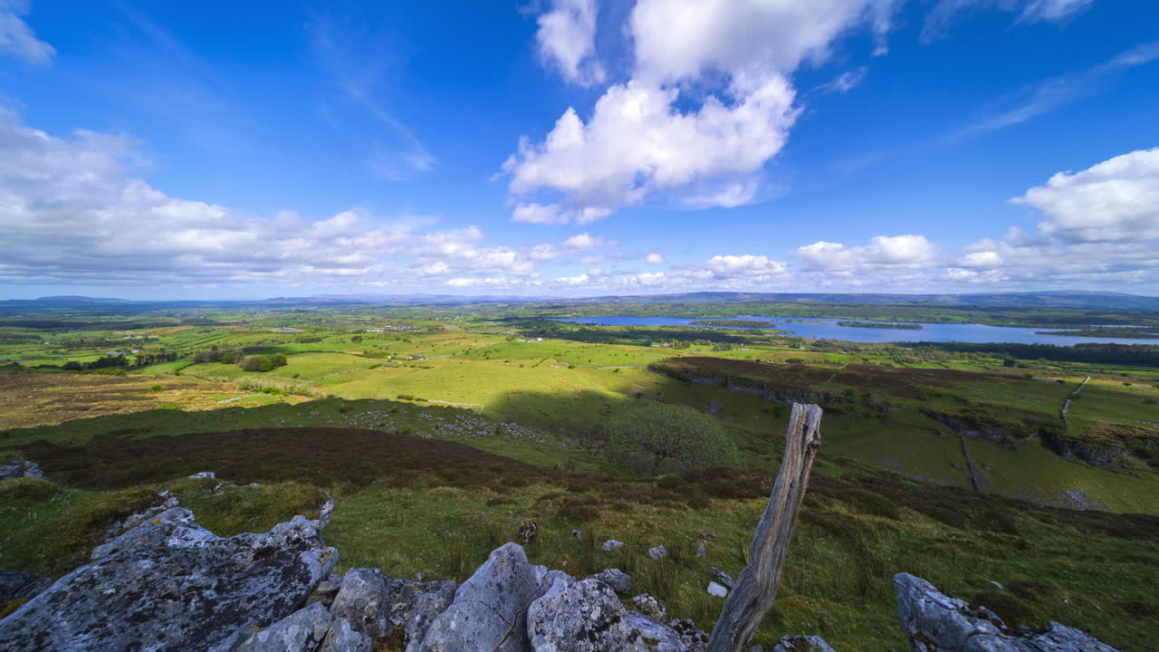 İrlanda, Sligo bölgesindeki Carrowkeel'den kayalık duvar ve ahşap sütunların ön planda olduğu, tepeler ve gölün uzakta bulunduğu güneşli ve bulutlu bir günde kırsal manzaranın panoramik hareketli hızlandırılmış çekimi