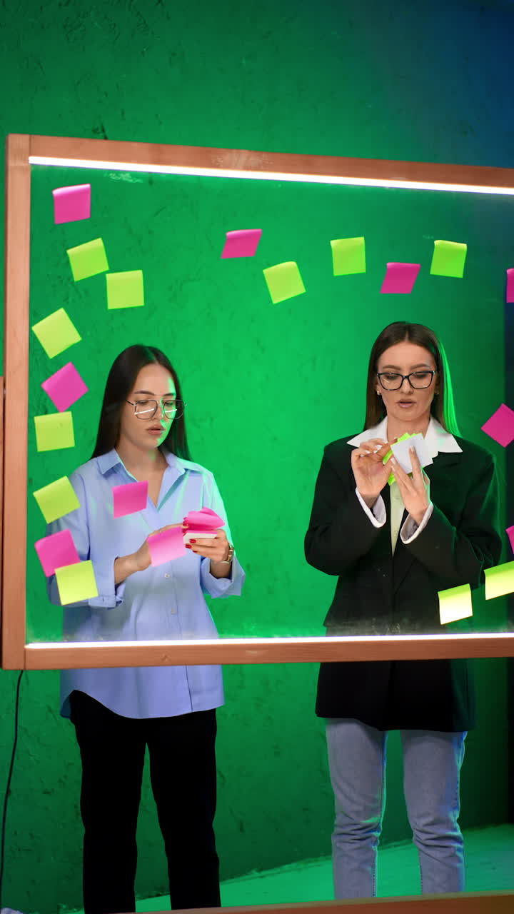 Two focused ladies stand behind the glass wall. Writing memos and putting them on the board for planning. Vertical video.