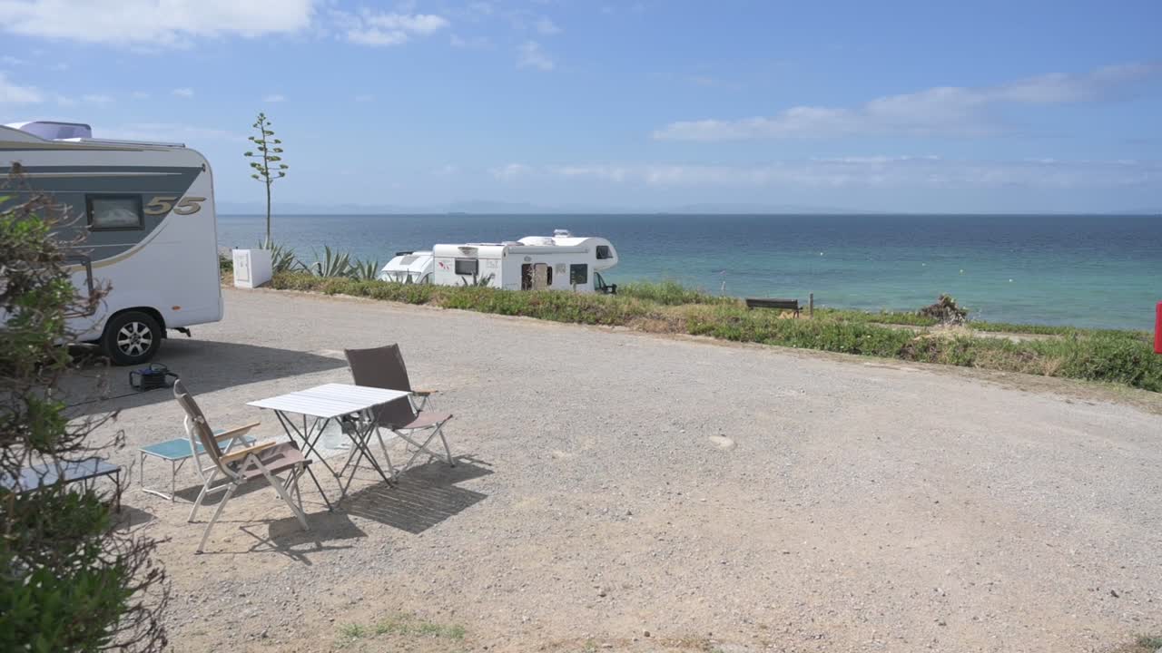Caravans parked in a designated area near the calm, blue sea on a sunny day. Table and chairs outside