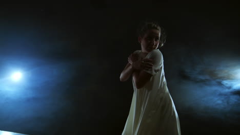 a young girl in a white dress dances contemporary in slow motion on the stage with smoke in spotlights. camera is moving around the dancer.