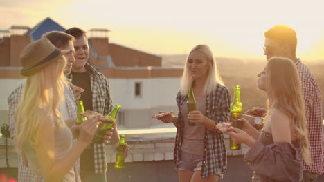 the company of six young people enjoy the time on the roof. they celebrate a friends birthday. they eat hot pizza and drink beer and enjoy smiles in the summer evening. they clink beer in the green bottles and communicate with each other.