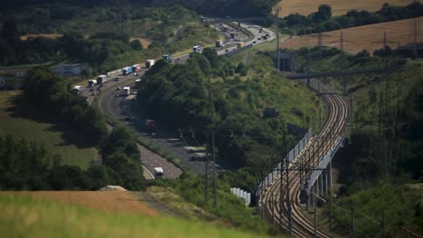 Lush-greenery-flanking-railway-and-highway-in-sunlight,-transportation-in-harmony-with-nature,-distant-perspective