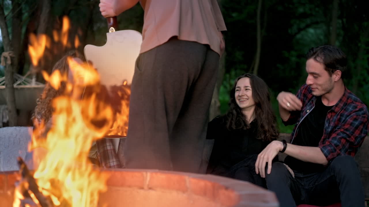 A group of happy young friends near a campfire at glamping, night. Man and women. Calling a guy with guitar. Slow motion