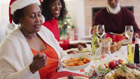 african american senior woman in santa hat serving food in plate while sitting on dining table havin