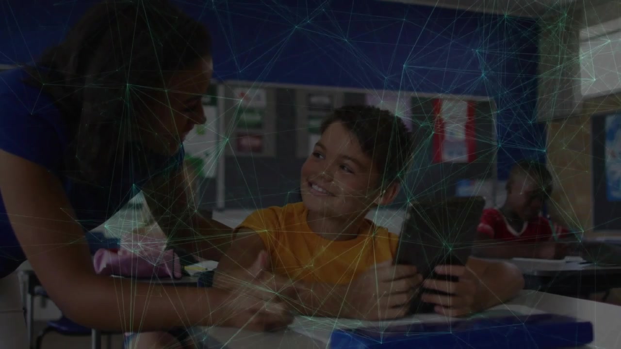 Boy holding tablet and smiling, teacher leaning in guiding pencil on worksheet for education