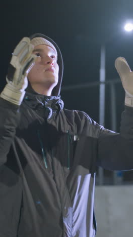 futbolista joven con guantes lanza una pelota de fútbol al aire durante la práctica nocturna en un campo urbano, el fondo incluye edificios altos, cercas de cadena y iluminación bokeh suave