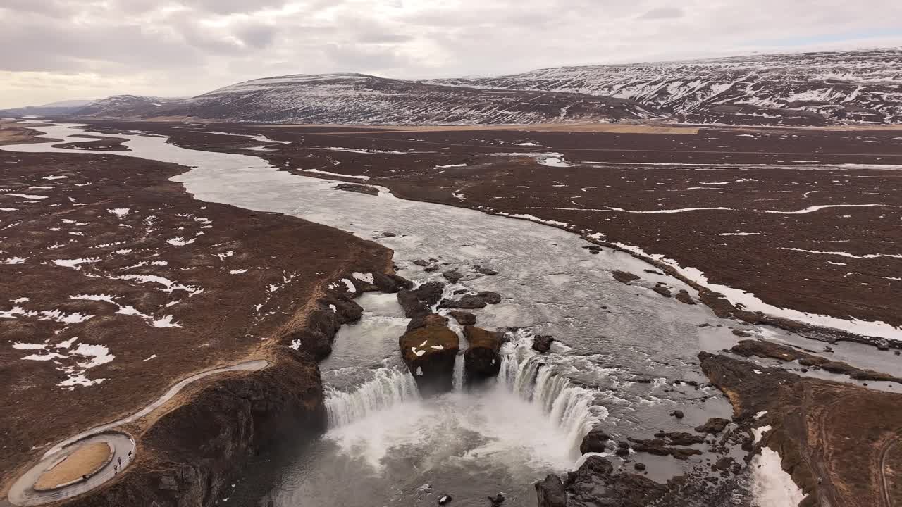 Aerial view of Goðafoss waterfall along the Skjálfandafljót river in Iceland, where icy waters plunge dramatically into a rugged canyon amid early spring thaw.
