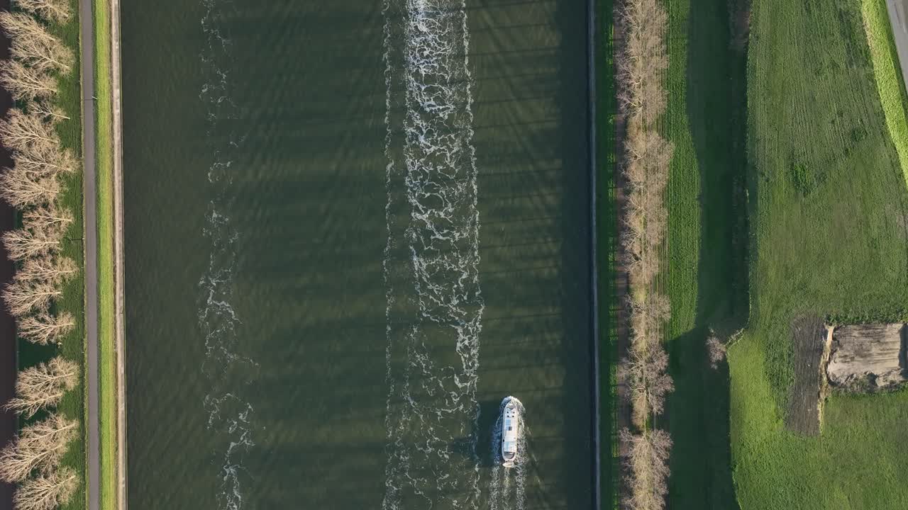 Top-down zenithal forward view of a small boat leaving wake lines on Antwerp canal, surrounded by trees and countryside fields