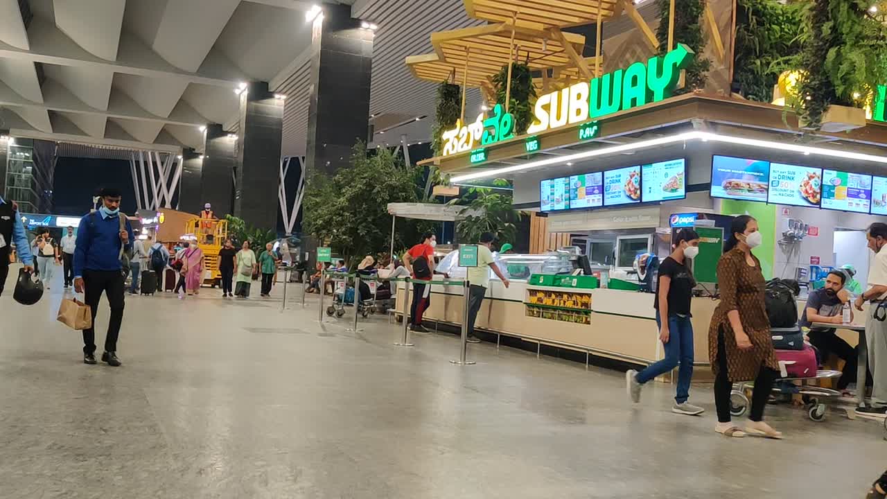 Bangalore airport passed next to the foodcourt with people standing waiting in line for the Subway sandwich store and passengers.