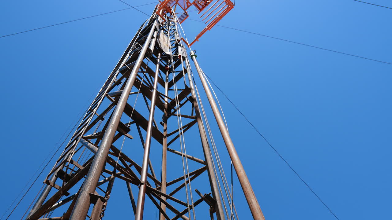 Long metal pipe attached to the wire is wobbling from side to side. A tower for drilling oil and gas beside. Low angle view.