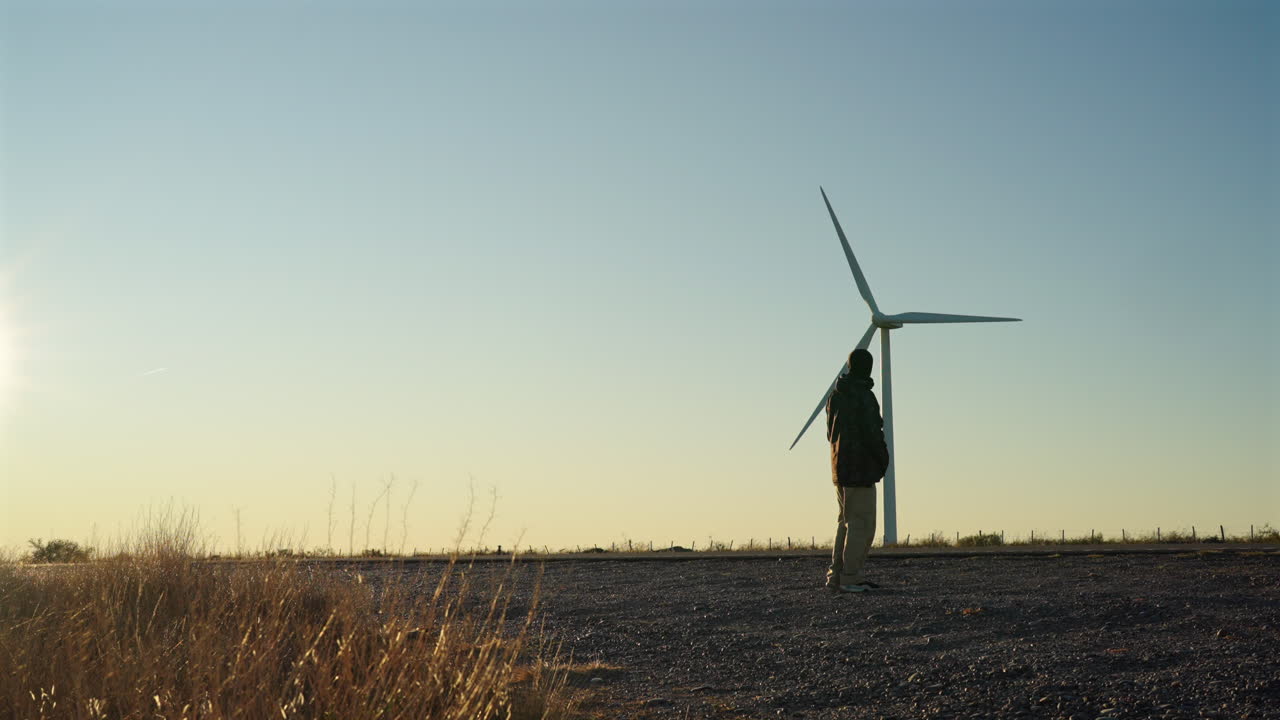 Man walks on a road near passing traffic under a wind turbine, then stops to watch the spinning blades, captured in real time with a static camera in golden light at sunrise in Patagonia Argentina