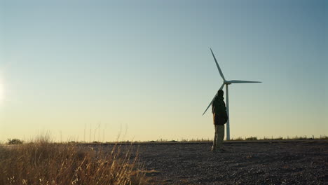 man walks on a road near passing traffic under a wind turbine, then stops to watch the spinning blades, captured in real time with a static camera in golden light at sunrise in patagonia argentina