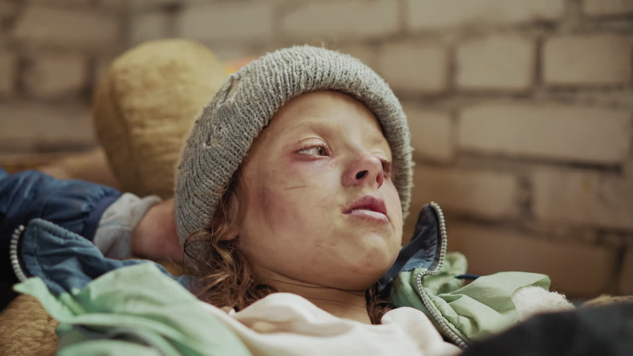 Close up of sick child with dirty face and worn clothes drinking water from metal cup while laying down, hand helping hold cup, capturing raw , struggle, and human compassion in harsh conditions