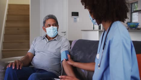 mixed race female doctor wearing mask and senior man taking blood pressure at home