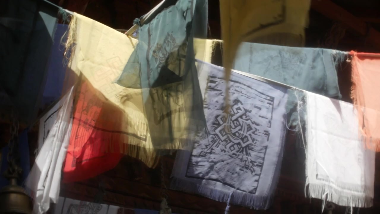 Drapeaux de prière flottant avec un léger flou de mouvement sous les avant-toits sombres d'un temple en bois. Une vue rapprochée montre un tissu à motifs, des chaînes suspendues, des ombres chaudes et une décoration culturelle.