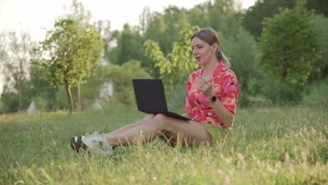 a middle-aged woman sits on the grass in the park. using a video link on a laptop, she conducts a conversation.