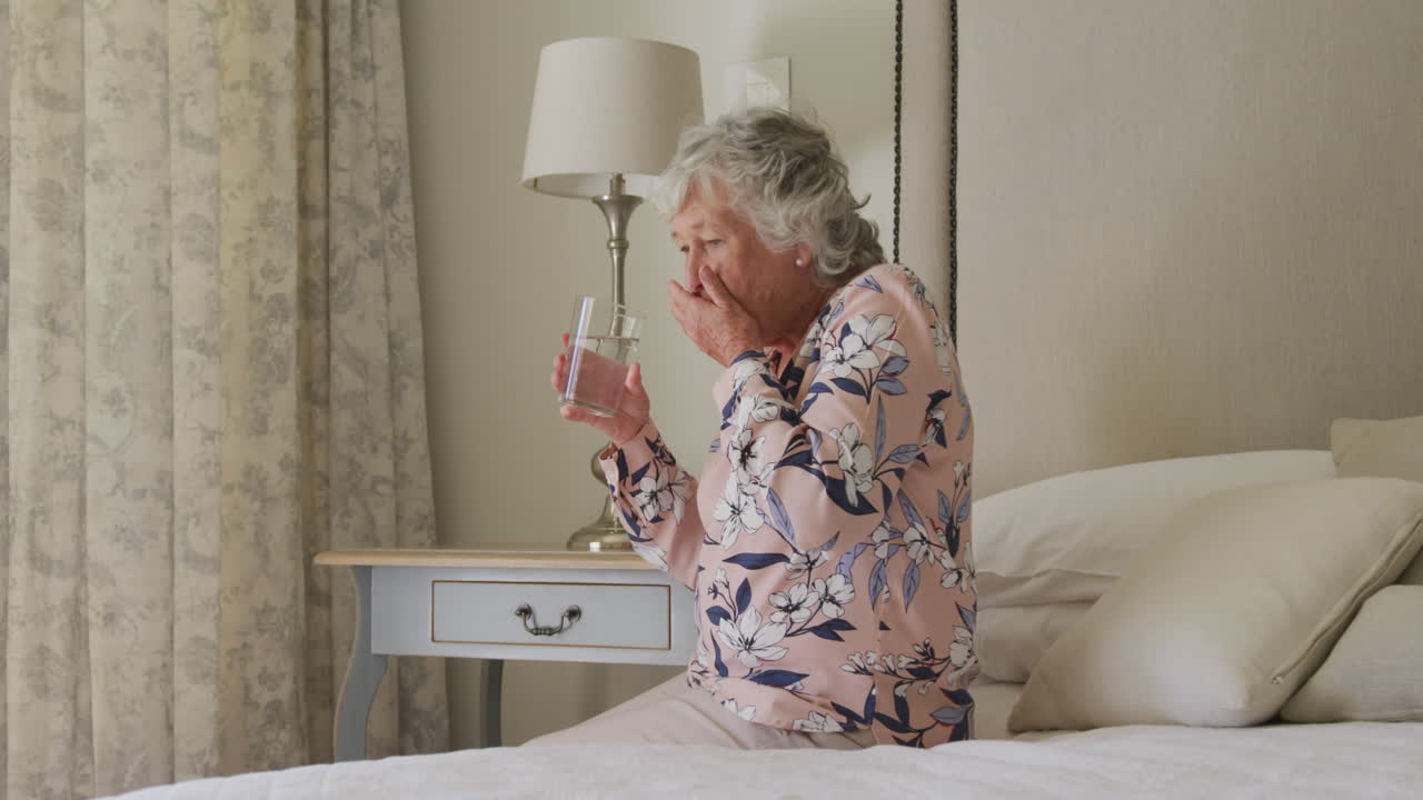 집에서 침대에 앉아서 의약품을 복용하는 백인 고령 여성의 중간 섹션