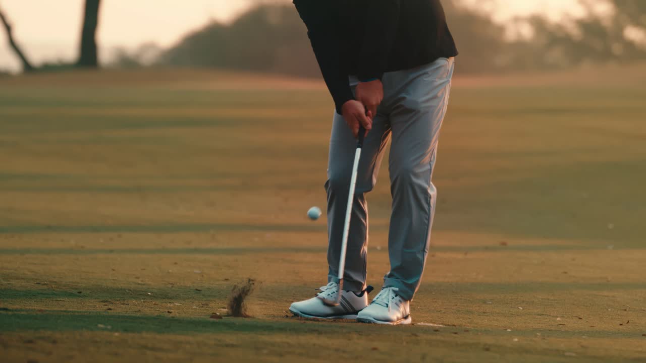 Rallentatore da angolazione bassa di un golfista che colpisce un wedge all'ora d'oro, con detriti di zolla che si sollevano su un campo da golf di un country club alberato. Ideale per pubblicità di marchi di golf, marketing di resort e campagne di abbigliamento