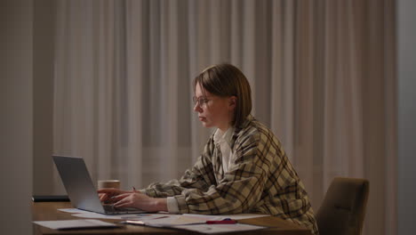 una chica trabaja desde casa o un estudiante está estudiando desde casa o un freelancer. ella usa una computadora portátil y un teléfono. la chica se sienta en el escritorio en la sala de estar estudia en la computadora portátil tomando notas concentrada mujer joven trabaja