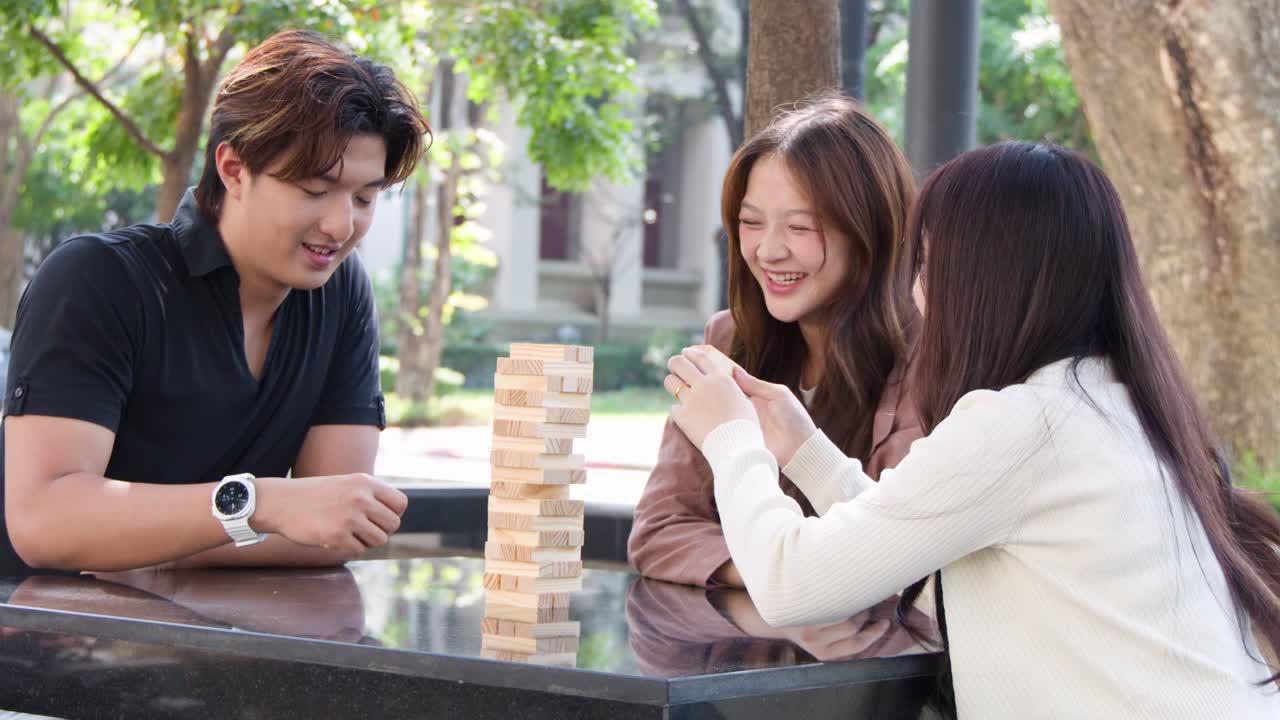กลุ่มเพื่อนวัยหนุ่มสาวกำลังหัวเราะขณะเล่นเกมตัวต่อไม้แบบหอคอยกลางแจ้งในเวลากลางวัน