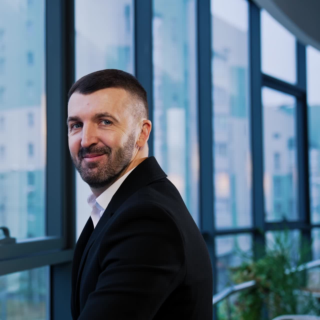 Good-looking bearded man stands at window and turns to camera. Businessman looking straight into camera smiling. Cityscape in blur at backdrop