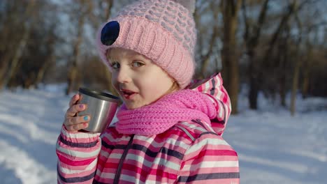 child girl kid drinking hot drink tea from cup on snowy road in winter park, christmas holidays