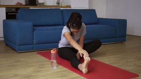 Young-african-woman-on-a-mat-in-living-room,-hard-to-do-sport-exercises,-doing-stretching