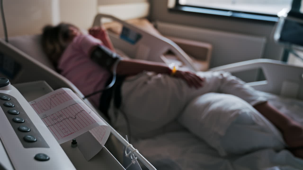 Close-up of a fetal monitor in a doctor's office, displaying fetal heart rate and uterine contraction data during a prenatal check-up with a blurred view of a pregnant woman lying in a hospital bed