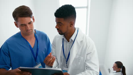 Male-Doctor-And-Nurse-With-Clipboard-Discussing-Patient-Notes-On-Stairs-In-Hospital-Building