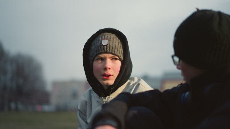Close-up of a young boy in a gray beanie and black hoodie, intently talking to another person in black, with a blur background