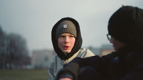 close-up of a young boy in a gray beanie and black hoodie, intently talking to another person in black, with a blur background
