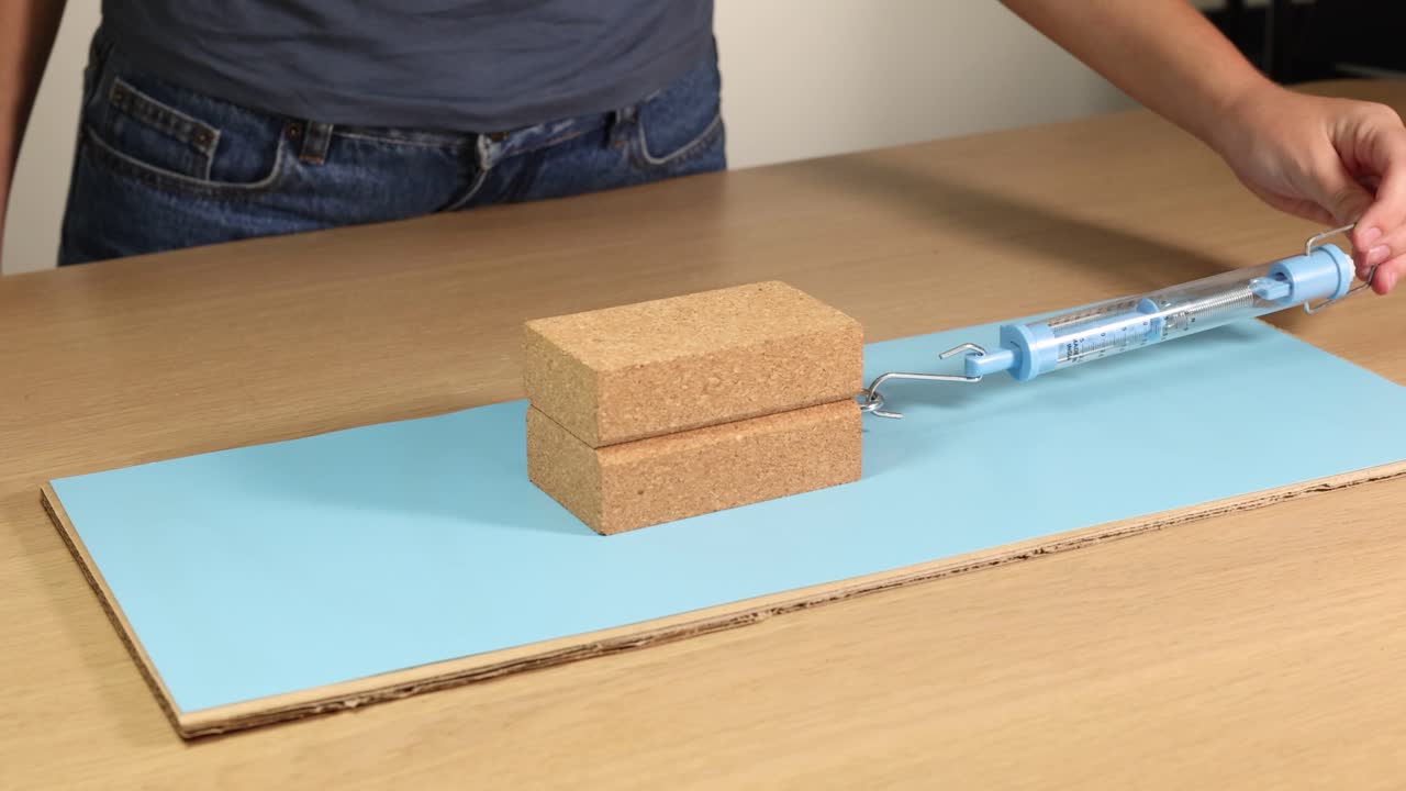 A wooden block is dragged across a surface using a spring scale, demonstrating friction and force measurement