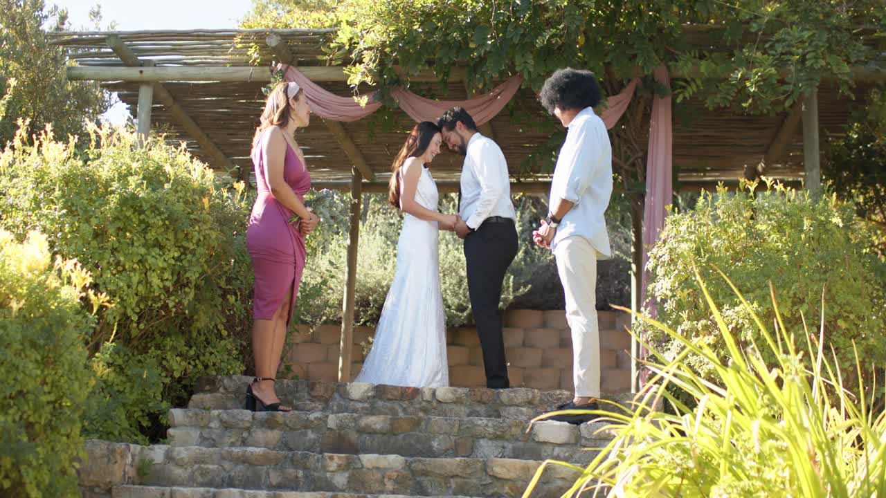 Pareja con atuendo de boda tomados de la mano, inclinando las cabezas durante los votos en escalones de piedra bajo una pérgola