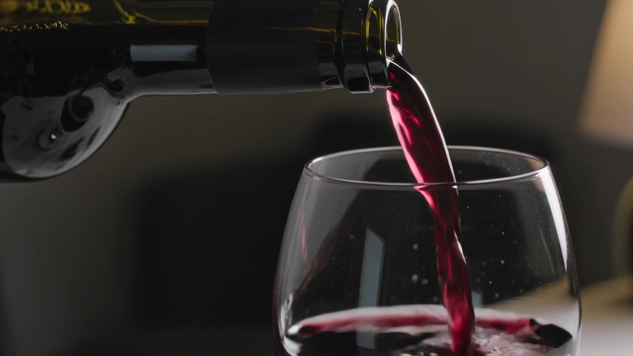 Botella de vino de cristal oscuro inclinada, vertiendo vino tinto en una copa sin tallo en casa para llenarla.