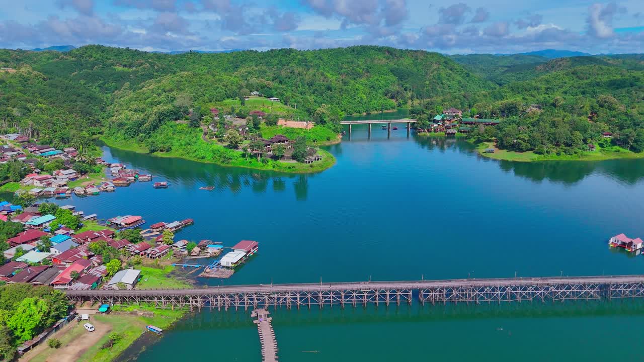 Aerial shot descending and tilting down over the Mon Bridge in Sangkhlaburi, Thailand. The long wooden structure spans calm blue water, with floating houses and lush green hills nearby