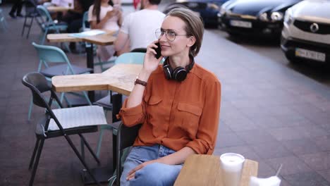 happy, confident blonde woman in denim and orange shirt sitting on a chair in a street cafe. talking by her smartphone. smiling