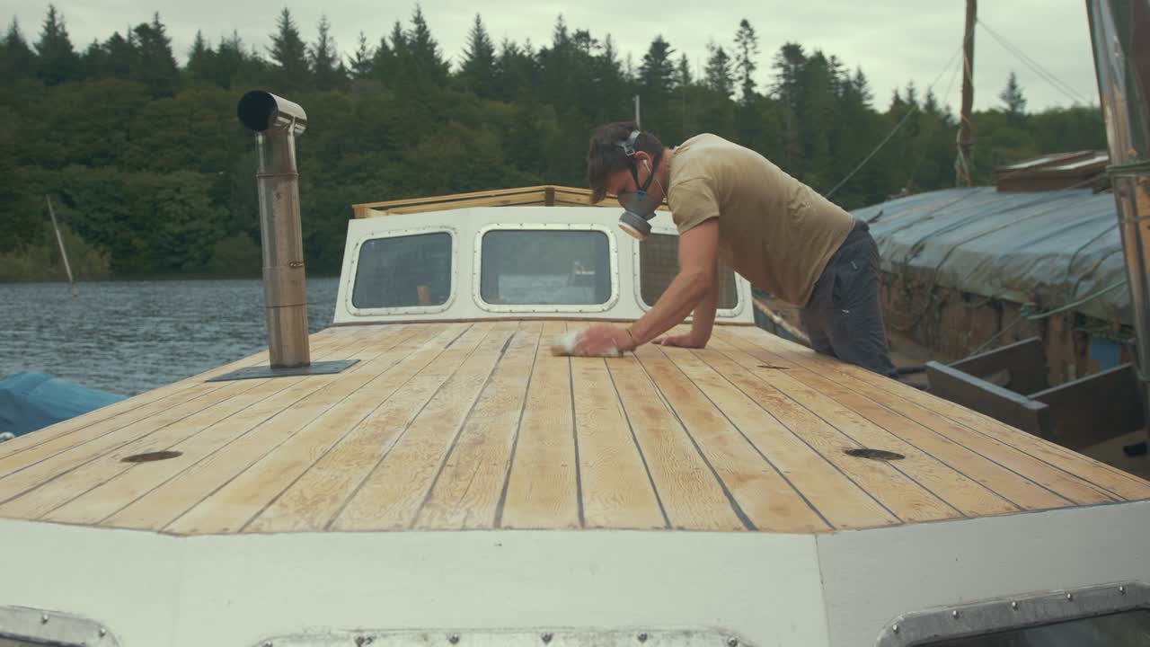 joven carpintero limpiando la cubierta lijada de un barco de madera a bordo