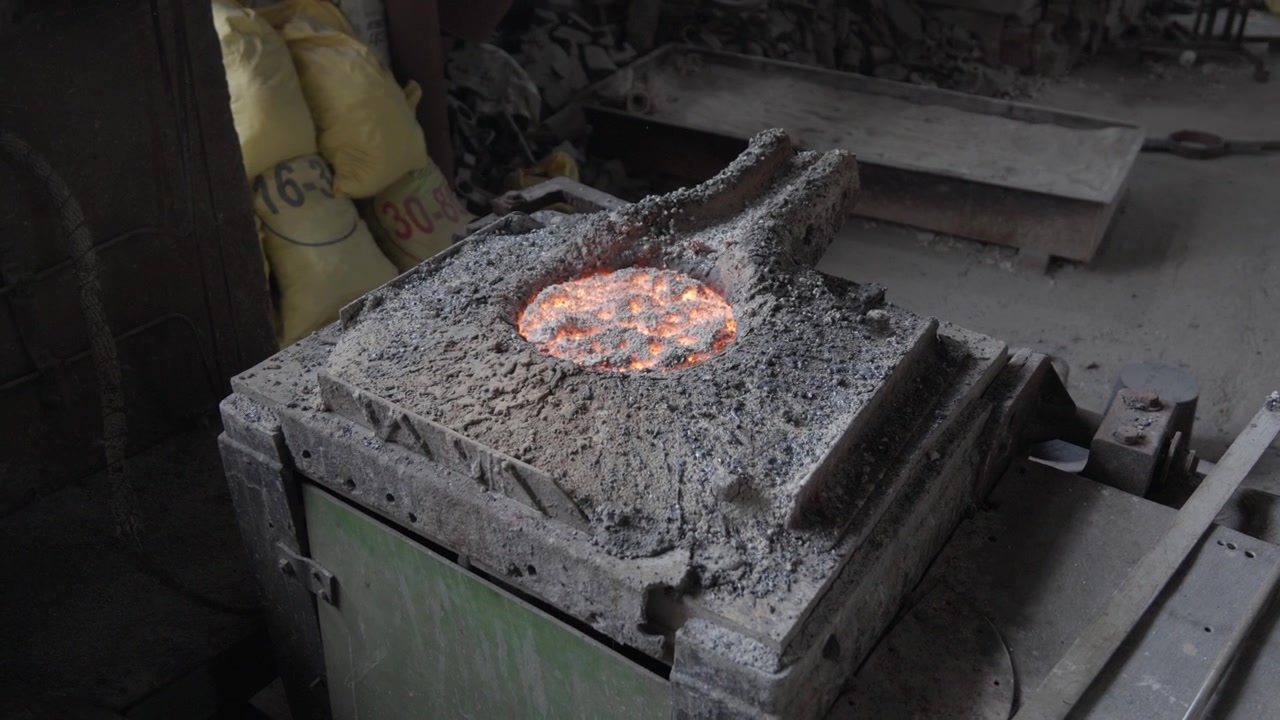 Un'apertura circolare di un forno rivela un bagno di metallo arancione parzialmente coperto da uno strato superficiale scuro all'interno di una cornice refrattaria grigia e ruvida
