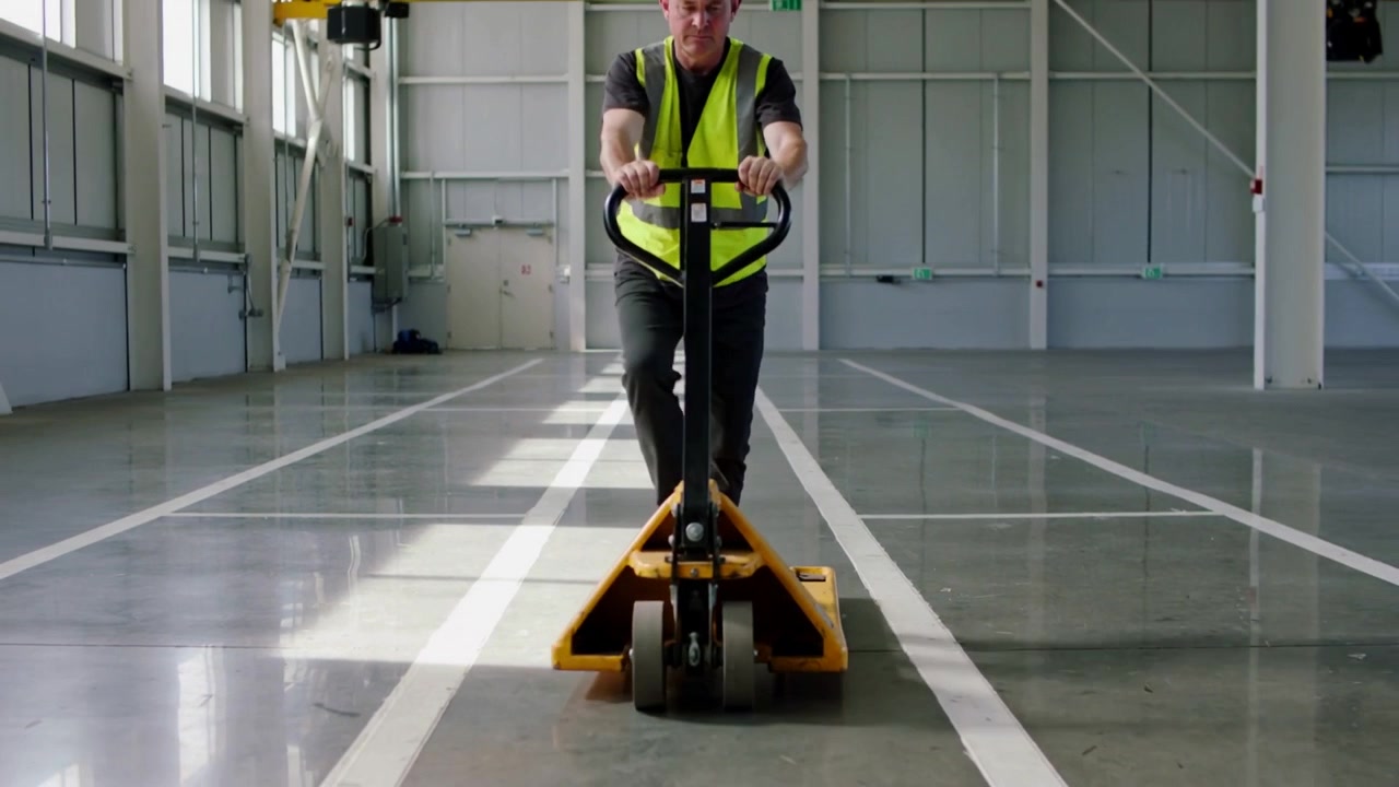 Pulling yellow pallet jack, worker spotting clear lane, moving in warehouse wearing vest hard hat