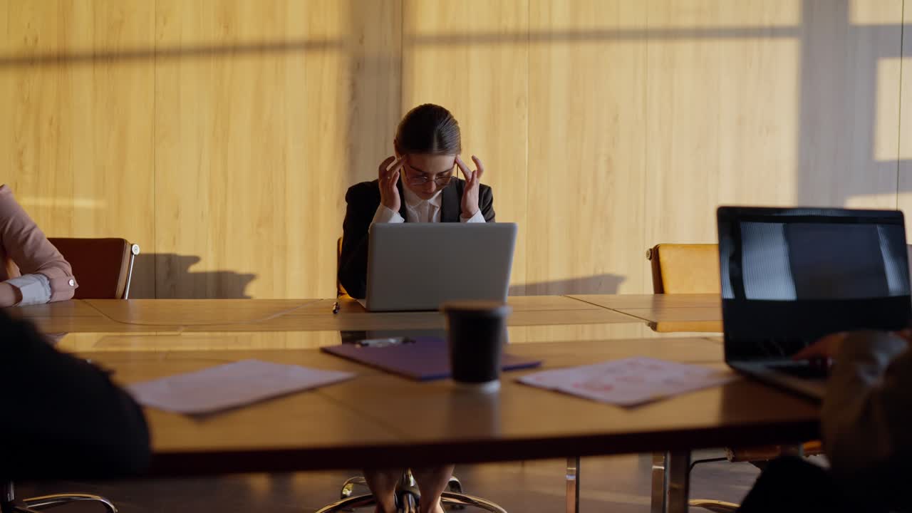 tired brunette girl in round glasses and a business suit sits at a wooden table in the office and massages her temples during a break in the tense atmosphere of the office