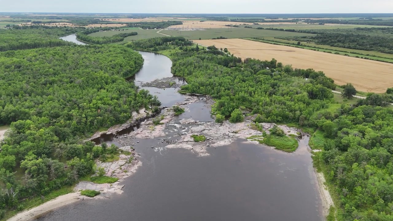 High aerial drone flight moving forward over Whitemouth Falls looking upstream along meandering Whitemouth River, surrounded by green boreal forest and farm fields in summer Manitoba.