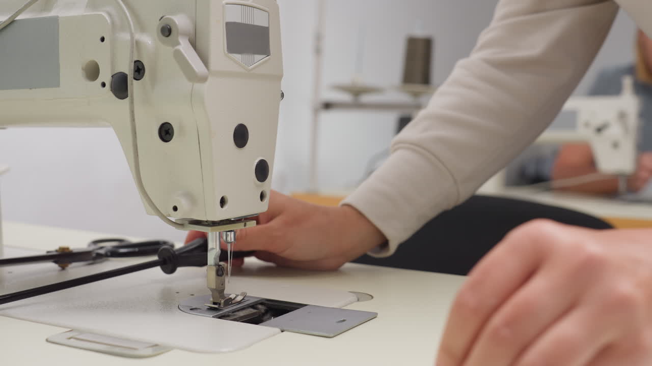 Woman seated adjusting sewing machine by loosening nut with screwdriver, working with focus in shared tailoring workspace as fabric and scissors lie on table beside her