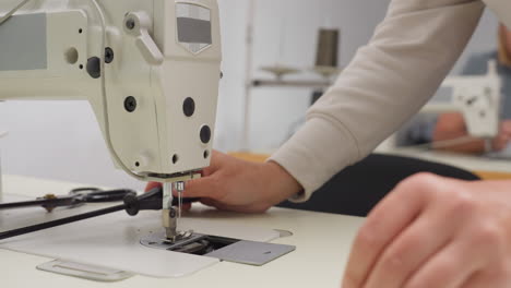 woman seated adjusting sewing machine by loosening nut with screwdriver, working with focus in shared tailoring workspace as fabric and scissors lie on table beside her