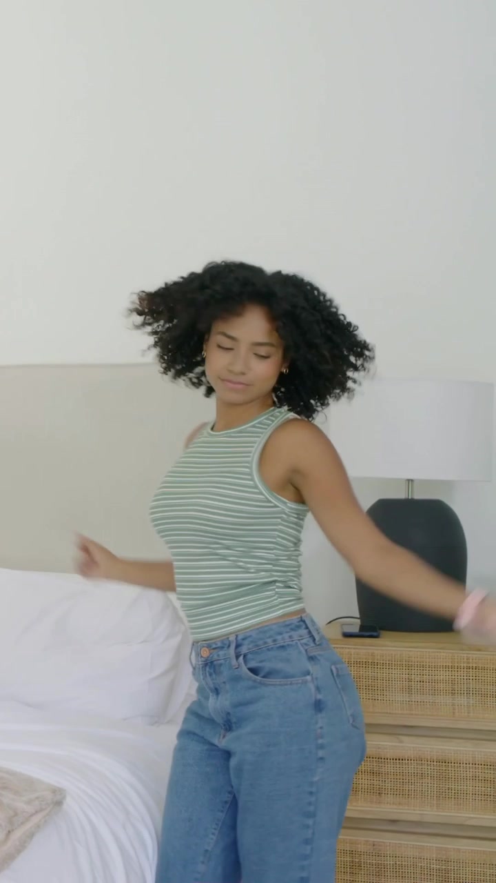 Video verticale: Una donna con una canotta verde esegue movimenti con le braccia accanto al letto, controllando il telefono.