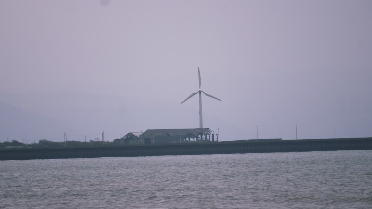 Pan Up to Single Small Wind Turbine Turning on Windy Day and Small Waves Roll into Swansea Bay with Port Nearby