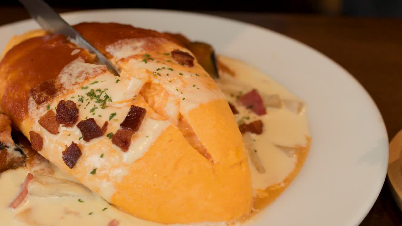 Knife cuts into omurice topped with creamy sauce and bacon in warm restaurant lighting