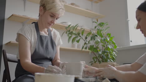 a female potter's wheel sculpting teacher explains how to work and teaches an elderly woman to work with clay and make mugs and jugs. master class for pensioners. pottery courses